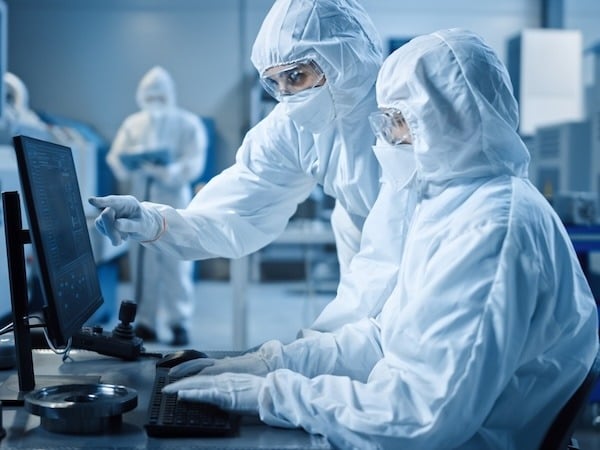Two workers wearing full-body cleanroom suits, gloves, face shields, and masks are operating a computer inside a sterile, high-tech laboratory or manufacturing cleanroom. One person is seated at the computer while the other points at the monitor. Additional personnel and equipment are visible in the background.
