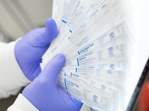 A person wearing purple gloves holds a fan of individually packaged sterile rayon-tipped swabs, displaying the product labels. The swabs are arranged against a bright white background, likely for inventory purposes.