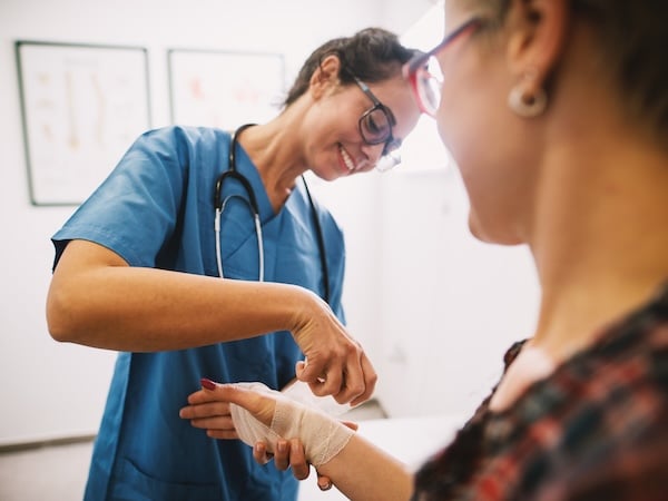 A healthcare professional in blue scrubs and glasses is smiling while wrapping a bandage around a patient's wrist. The patient, who is wearing glasses and a plaid shirt, looks on.