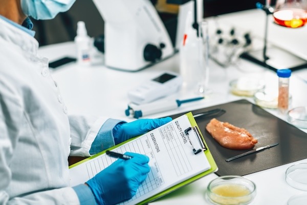 A food safety technician in a laboratory, wearing a white lab coat and blue gloves, writes on a quality control form on a clipboard. In the background, there are laboratory instruments and a sample of chicken meat on a black cutting board, alongside other samples and test equipment on a white lab bench.