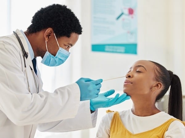 A healthcare professional wearing a mask and gloves performs a nasal swab test on a young woman. The woman, seated and relaxed, tilts her head slightly back as the swab is gently inserted into her nostril. The background shows a clinical setting with a poster on the wall.