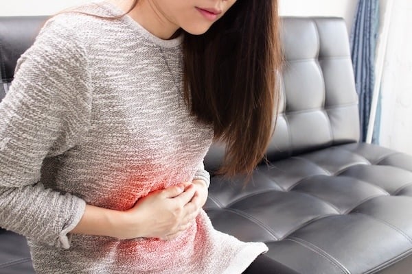 A woman sitting on a black leather couch, clutching her stomach in discomfort. A red overlay highlights the abdominal area, indicating pain or digestive distress.