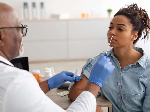 A healthcare professional wearing blue gloves and glasses is administering a nasal swab test to a young woman with curly hair, who is seated and wearing a denim shirt.