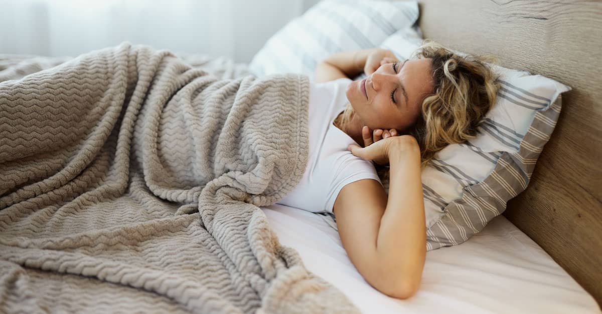 Woman sleeping peacefully under a soft beige blanket in a cozy winter bedroom, showing comfort, warmth, and relaxation for better winter sleep.