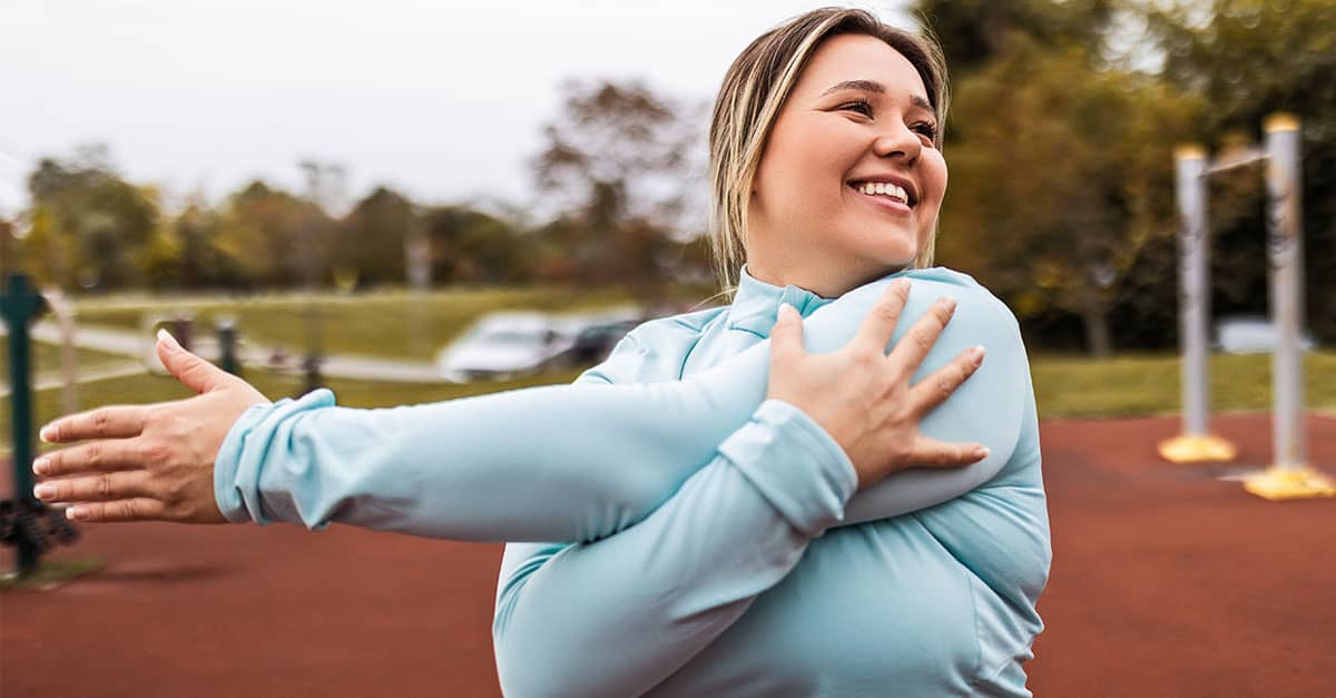 Smiling woman doing an upper body warm up stretch outdoors, showing dynamic vs static stretching to loosen her shoulders before exercise.