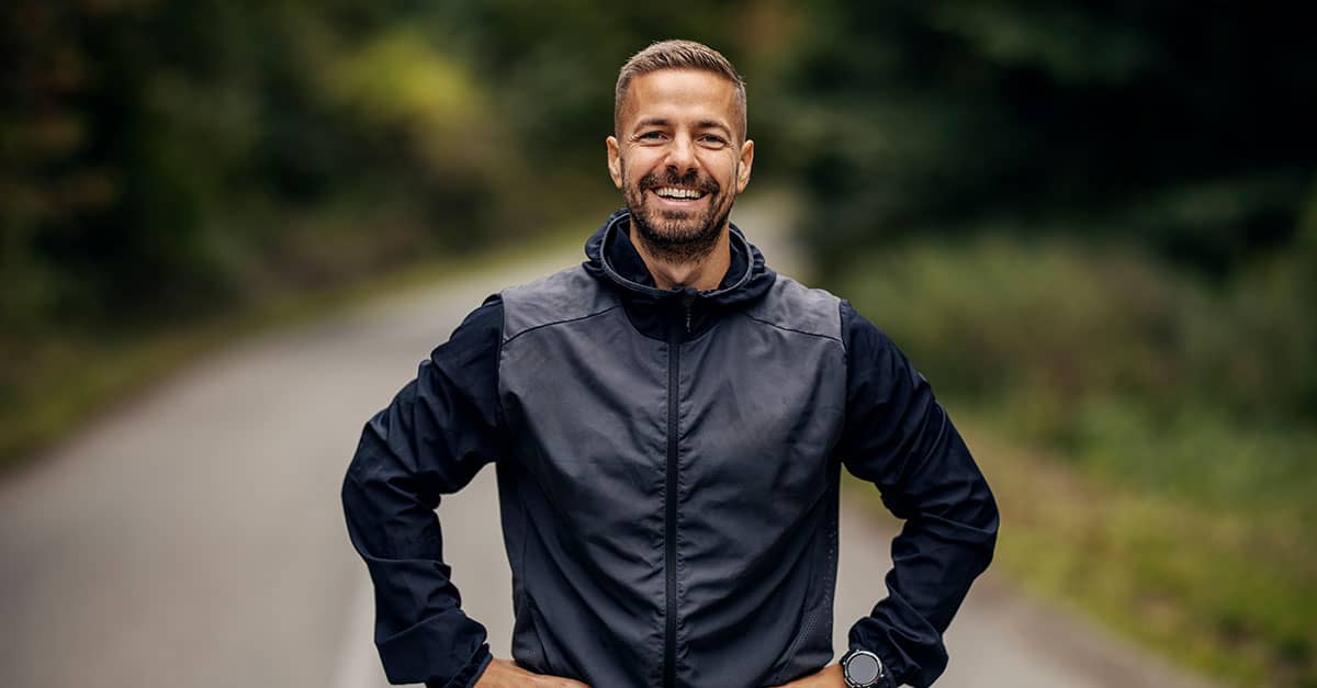 Smiling man standing tall with hands on hips, demonstrating what good posture looks like on an outdoor road.